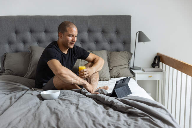 Вид сбоку восхищенного мужчины, сидящего на кровати со стаканом апельсинового сока и просматривающего таблицу, завтракая утром дома — стоковое фото