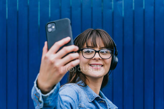 Mujer hipster feliz en los auriculares tomando auto disparo en el teléfono móvil en el fondo de la pared azul en la calle - foto de stock