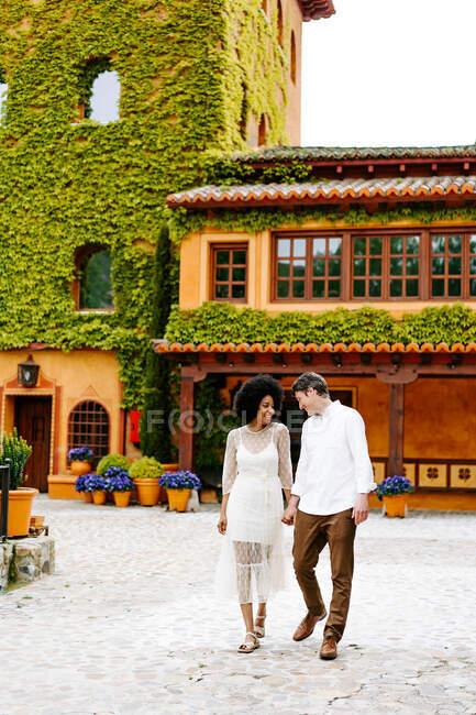 Loving man holding hands of black woman while standing near building with ivy on walls in park in summer — Stock Photo