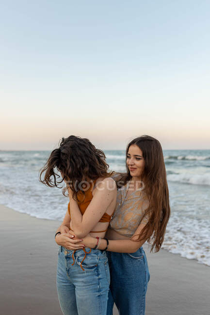 Jovens namoradas abraçando uns aos outros enquanto estão de pé na praia de areia perto do mar acenando ao pôr do sol — Fotografia de Stock