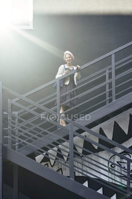 Délicieuse femelle en robe debout sur un escalier noir éclairé par la lumière du soleil et regardant la caméra — Photo de stock
