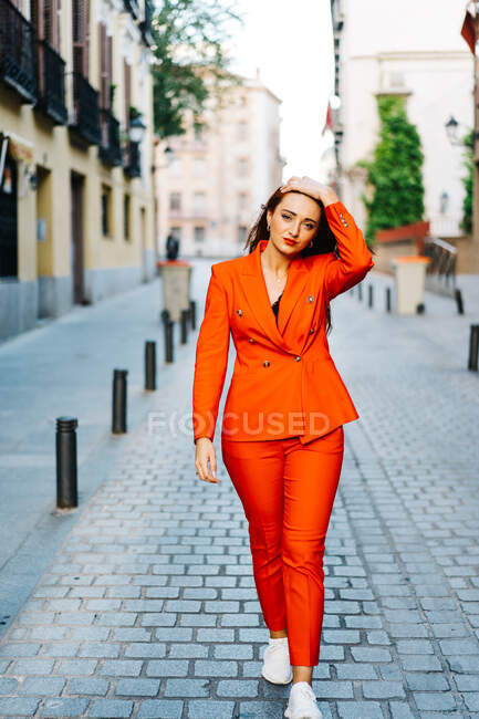 Donna determinata in elegante abito arancione brillante toccare i capelli lunghi zenzero e camminare lungo la strada in città mentre guardando la fotocamera — Foto stock