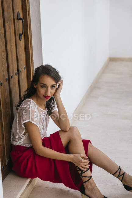 Seitenansicht einer fröhlichen ethnischen Frau mit roten Lippen und in trendiger Sommerkleidung, die neben einer Holztür im Innenhof des Hauses sitzt und wegschaut — Stockfoto