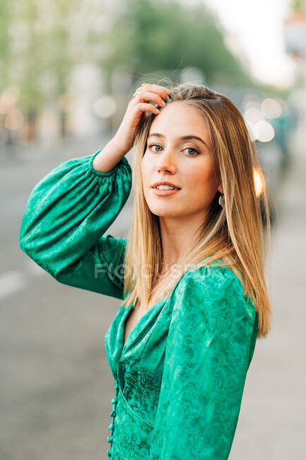 Donna spensierata in abito verde alla moda in piedi con un braccio alzato e toccando la testa in strada e guardando la fotocamera — Foto stock