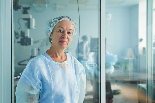 Amable médico de mediana edad en uniforme quirúrgico mirando a la cámara contra colega irreconocible en el hospital - foto de stock