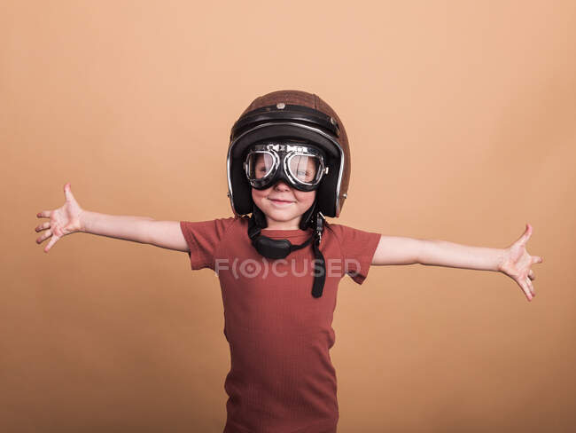 Весела дитина в шоломі і захисні окуляри дивиться на камеру з розтягнутими руками на бежевому фоні — стокове фото