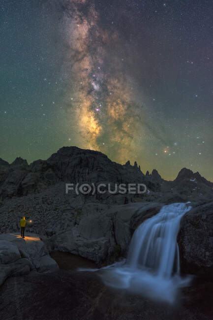 Анонимный путешественник с факелом, созерцающий водопад, струящийся среди неровной скалистой местности под ночным звездным небом с ярким светящимся Млечным Путем — стоковое фото