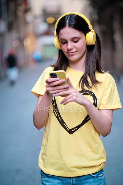 Stylish female in yellow headphones and t shirt listening to songs while standing browsing smartphone in city street — Stock Photo