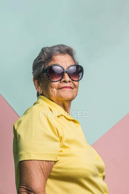 Heureuse femme âgée moderne aux cheveux gris et aux lunettes de soleil tendance sur fond rose en studio et en regardant la caméra — Photo de stock