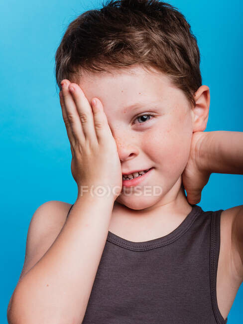 Shy cute preteen boy covering eye with hand and looking at camera on vivid blue background in studio — Stock Photo