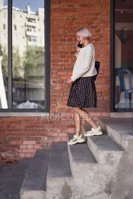 Side view of delighted female in trendy outfit walking downstairs in city and having conversation on mobile phone — Stock Photo