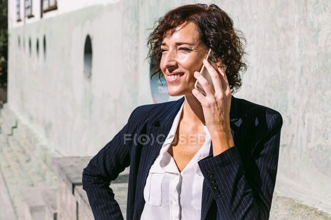Operaia esecutiva adulta positiva che indossa un vestito di classe in piedi con mano sulla vita e parla al cellulare nella giornata di sole — Foto stock