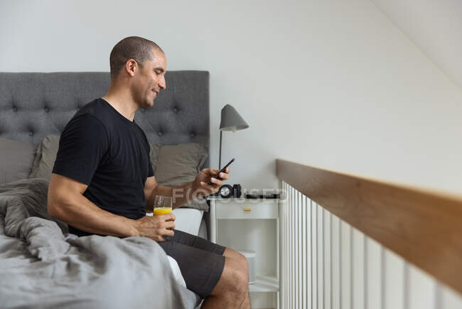 Vista lateral del hombre sentado en la cama de navegación en el teléfono móvil durante el desayuno en la mañana en casa - foto de stock