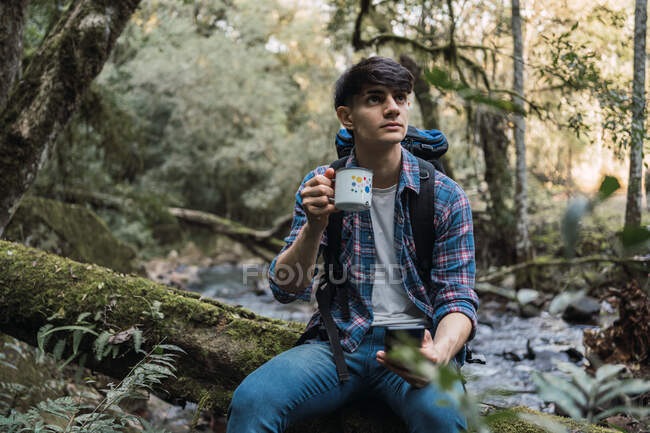 Friedlicher männlicher Entdecker mit Rucksack sitzt auf Baumstamm im Wald und trinkt während des Trekkings Heißgetränk aus Metallbecher — Stockfoto