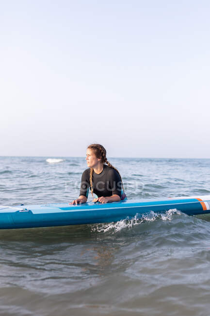 Female surfer lying on SUP board and floating on calm water of sea on sunny day looking away — Stock Photo