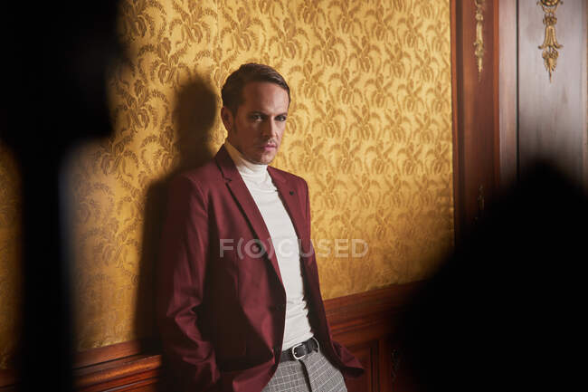 Уверенный взрослый актер в элегантной стильной одежде, держа руку в кармане и вдумчиво глядя в сторону, стоя у стены в винтажном стиле комнате — стоковое фото