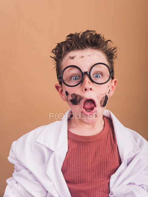 Niño sorprendido con la cara sucia y el pelo sin peinar en gafas decorativas mirando a la cámara sobre fondo beige - foto de stock
