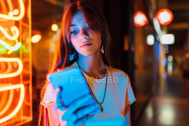 Цікава молода жінка в сережках, що серфінгують Інтернет на мобільному телефоні проти блискучих неонових ламп в місті — стокове фото