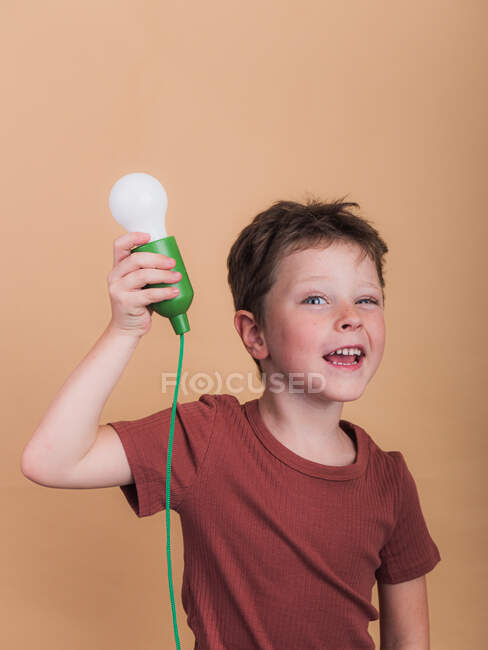 Дивовижна дитина в сорочці з пластиковою лампочкою, що представляє ідею, дивлячись на камеру на бежевому фоні — стокове фото