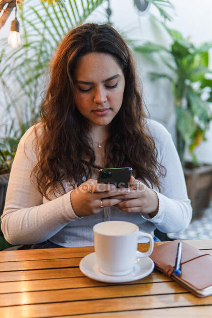 Young ethnic female messaging on mobile phone while sitting at table with cup of coffee and notebook in cafeteria — Stock Photo