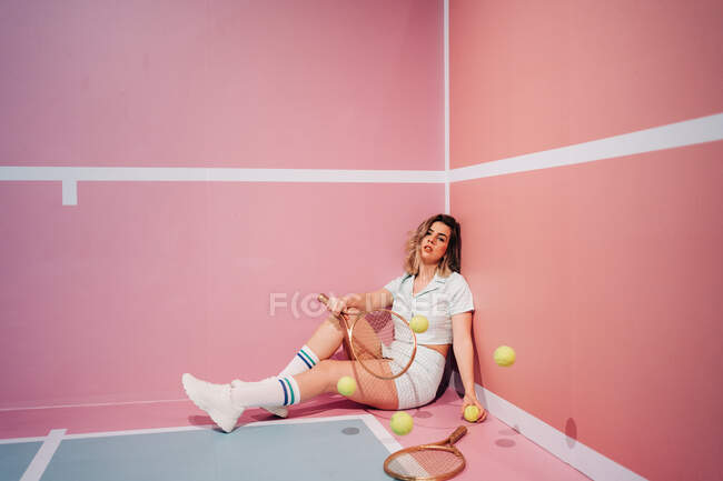Young sportswoman in sneakers and sports clothes sitting with tennis rackets while looking at camera against moving balls — Stock Photo