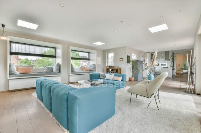 Diseño interior de sala de estar de espacio abierto con sofá azul y sillas colocadas cerca de una pequeña mesa en una alfombra suave en un apartamento moderno con paredes blancas y techo iluminado con lámparas - foto de stock