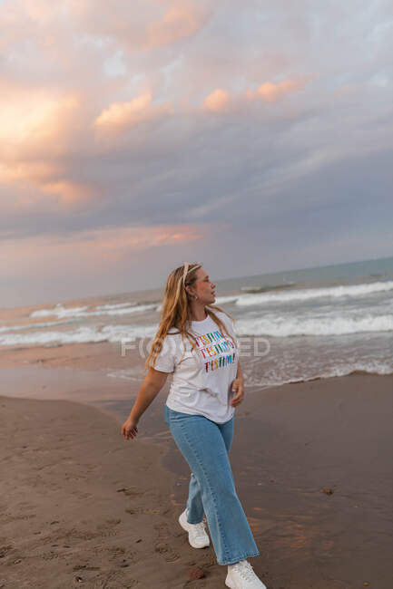 Mujer joven positiva en gafas de sol de moda y ropa elegante caminando en la playa contra el mar en la noche de verano - foto de stock