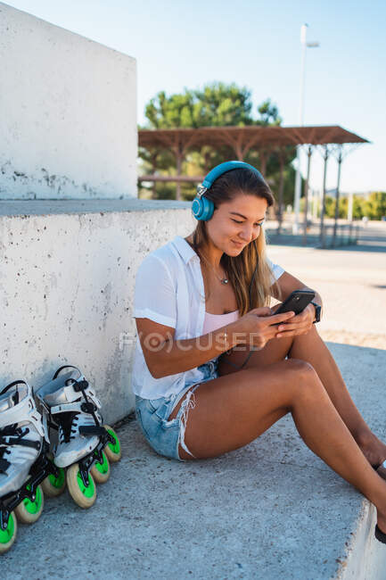Side view of positive cheerful young female browsing on mobile phone listening music on headphones on sunny day in summer in city — Stock Photo