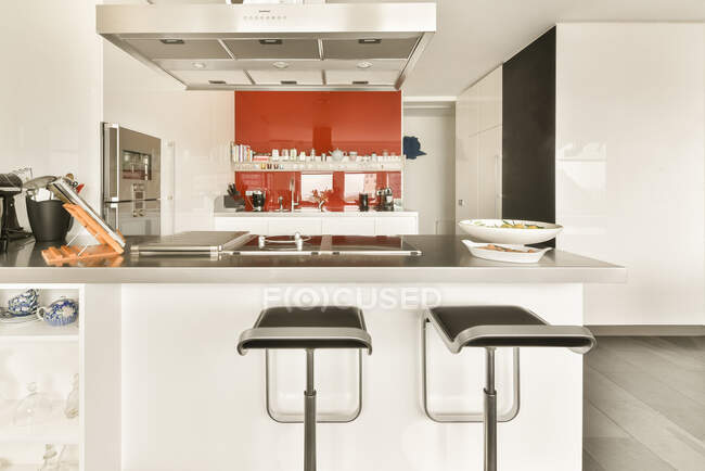 Kücheninsel mit Theke und Barhockern unter der Haube in moderner Freiraumwohnung mit weißen Wänden mit Möbeln und Utensilien — Stockfoto