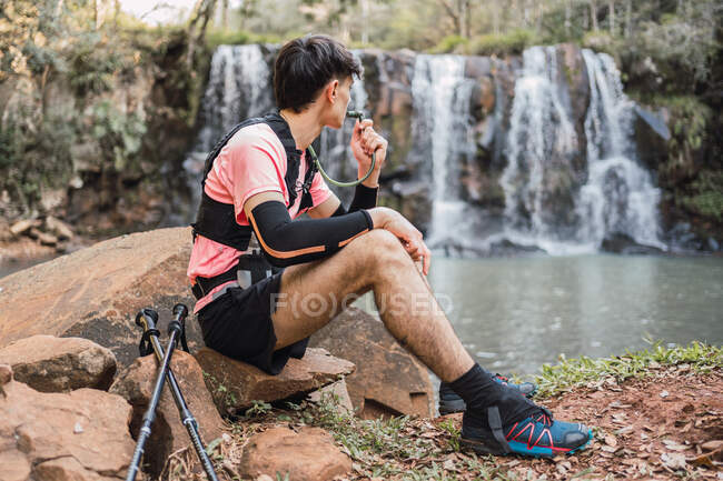 Вид збоку чоловічого пішохода з гідратаційним пакетом, що п'є від шланга, під час походу в ліс і захоплюючий вид на озеро біля водоспаду — стокове фото