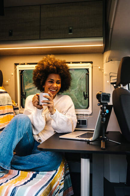 Cheerful young African American female traveler drinking coffee and enjoying video chat via laptop while resting inside camper van during summer adventure — Stock Photo