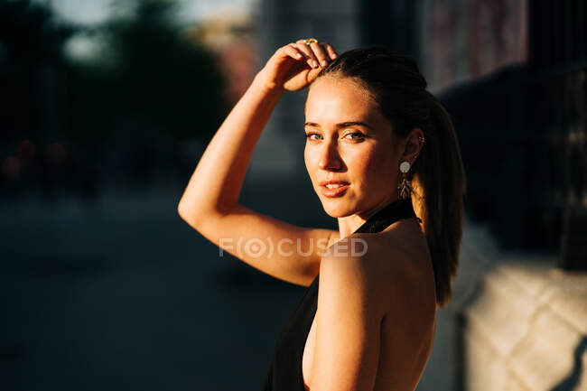 Vista lateral da mulher feliz vestindo roupas da moda em pé na cidade no dia ensolarado e olhando para a câmera — Fotografia de Stock