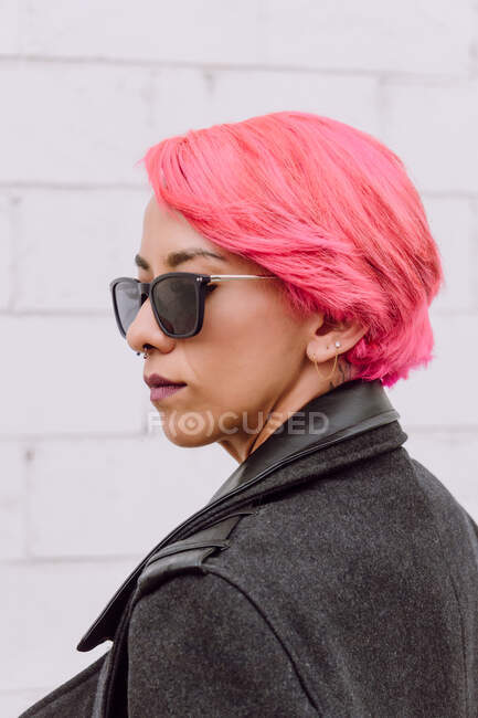 Stylisches weibliches Model mit pinkfarbenen Haaren im modischen Mantel und Sonnenbrille, die an der weißen Backsteinwand wegschaut — Stockfoto