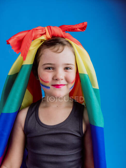Petite fille avec un drapeau arc-en-ciel attaché sur la tête regardant la caméra tout en se tenant debout sur fond bleu — Photo de stock