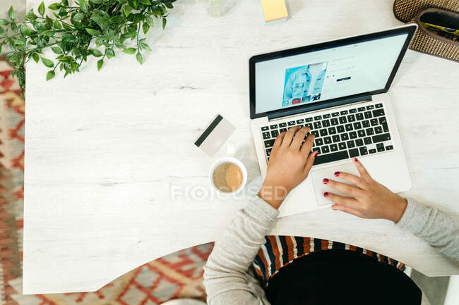 De dessus de la femme en utilisant netbook pendant les achats en ligne tout en étant assis à table avec une carte en plastique à la maison et en choisissant des marchandises — Photo de stock
