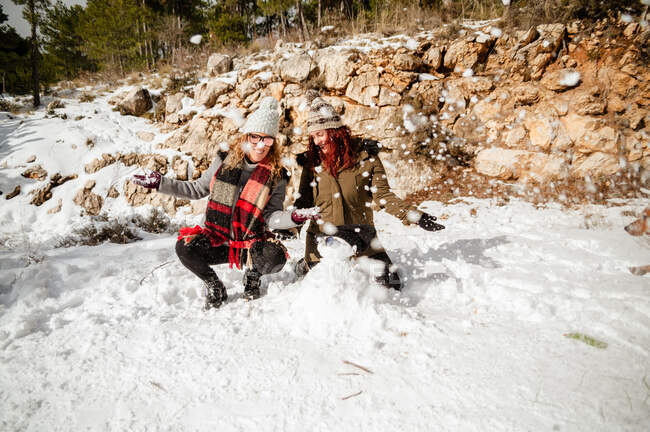 Entzückte Freundinnen in Oberbekleidung werfen Schnee und amüsieren sich an sonnigen Tagen im Winterwald — Stockfoto