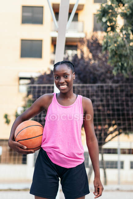 Cheerful African American female in activewear standing with ball on basketball playground and looking at camera — Stock Photo