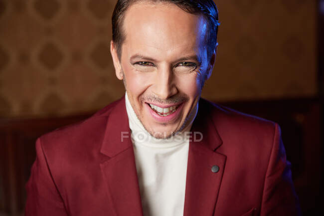 Elegante artista de teatro masculino adulto alegre en chaqueta de color burdeos y con maquillaje mirando a la cámara sonriendo - foto de stock