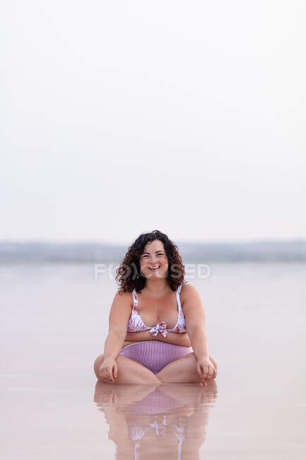 Delighted curvy female in bikini sitting in water of pink pond in summer and looking at camera — Stock Photo