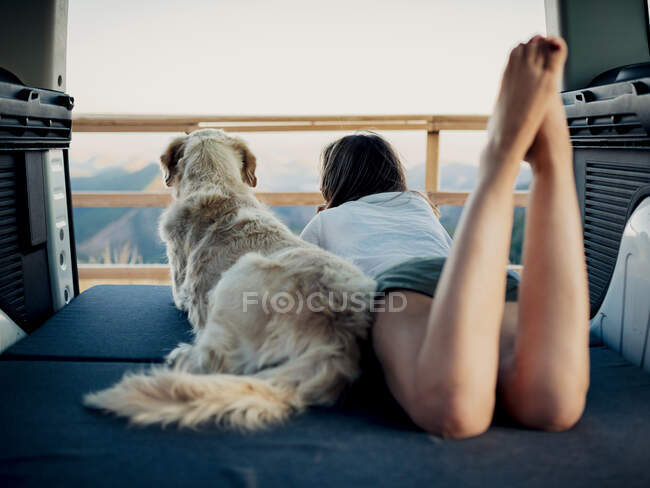Unrecognizable female tourist and obedient Golden Retriever dog lying on mattress inside camper and admiring nature — Stock Photo