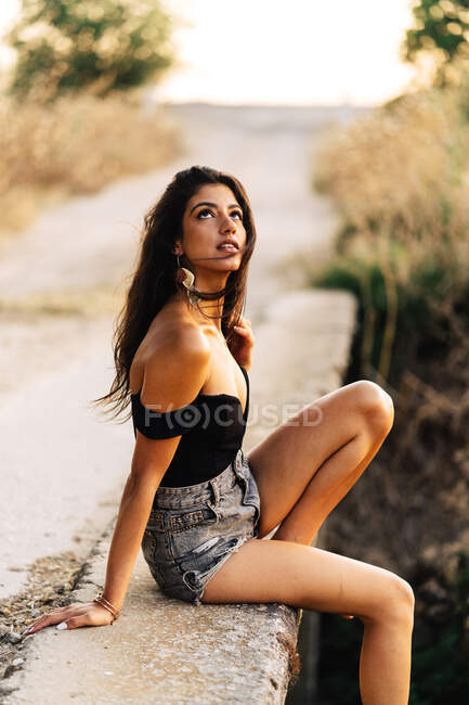 Vista lateral de la joven hispana descalza descalza en top negro y pantalones cortos de mezclilla grises sentados en puente de piedra en el día de verano en el campo - foto de stock