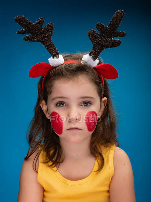 Menina séria com bochechas pintadas de vermelho usando chifres de veado de brinquedo e orelhas e olhando para a câmera no fundo azul — Fotografia de Stock