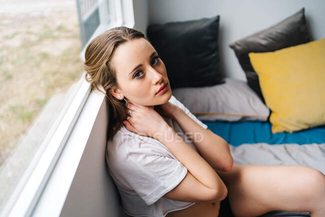 Вид сбоку деликатной женщины, трогающей шею, сидя на мягкой кровати у окна дома и глядя в камеру — стоковое фото