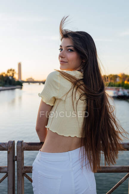 Vista posteriore della giovane femmina gentile che guarda la fotocamera contro la recinzione sul ponte in luce solare morbida — Foto stock