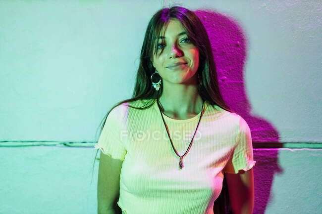 Очаровательная молодая женщина с длинными волосами смотрит в камеру, прислонившись к стене в вечернем городе — стоковое фото