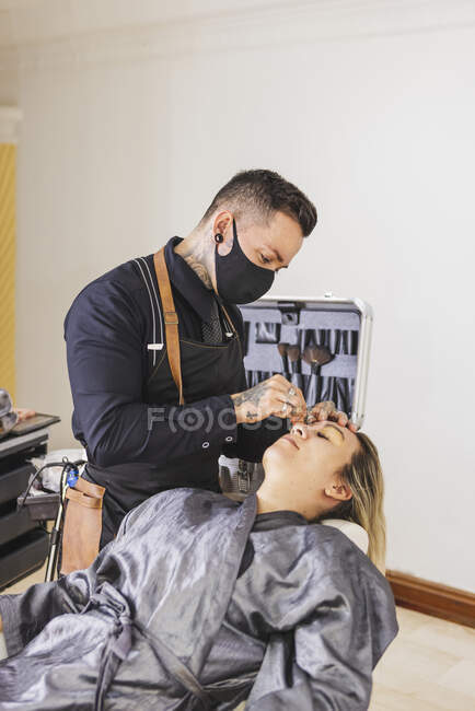 Mann mit Maske beschmiert Gesicht einer blonden Frau bei Arbeit im professionellen Make-up-Studio — Stockfoto