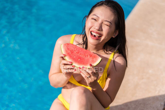 Von oben eine fröhliche ethnische Frau im gelben Bikini, die in der Nähe des Swimmingpools sitzt und an einem sonnigen Sommertag frische Wassermelone isst, während sie in die Kamera schaut — Stockfoto