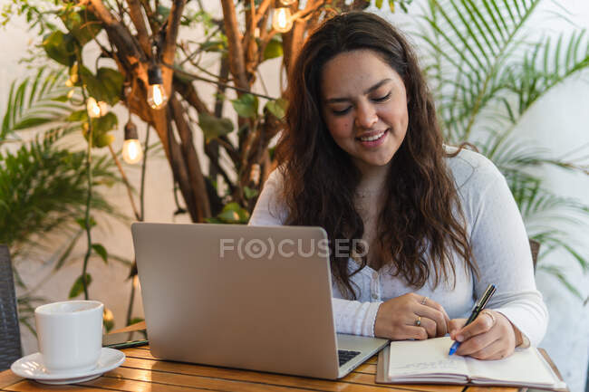Focalisé jeune étudiante latino-américaine prenant des notes dans le planificateur tout en travaillant sur ordinateur portable se préparant pour l'examen universitaire dans un café confortable — Photo de stock