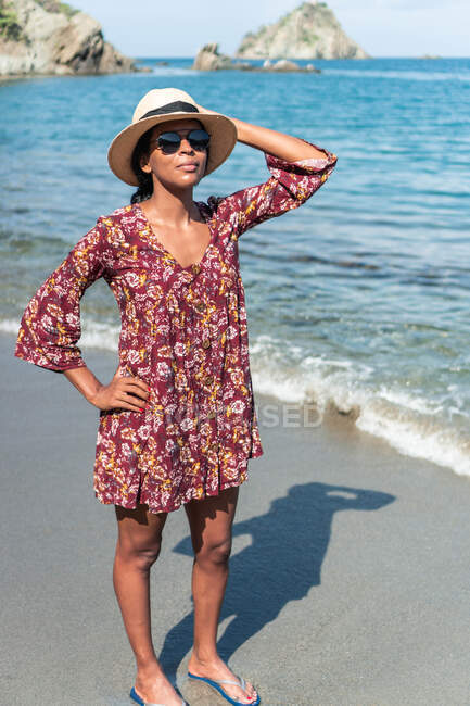 Ethnic female tourist in sundress standing with hand on hip on sandy coast against ocean and mounts in sunlight — Stock Photo