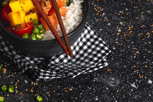 Высокий угол азиатского тычка с лососем и рисом с разнообразными овощами подается в миске на столе с палочками в ресторане — стоковое фото